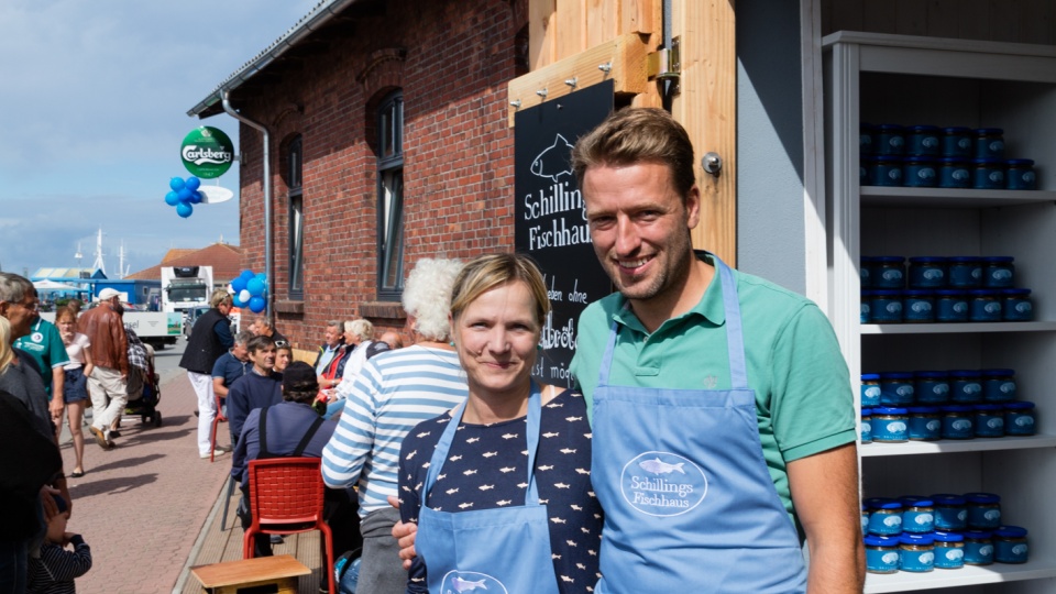 Nicolle und Matthias Schilling stehen vor dem Schillings Fischhaus in Schaprode auf Rügen.Sie tragen blaue Schürzen mit dem Fischhaus-Logo.